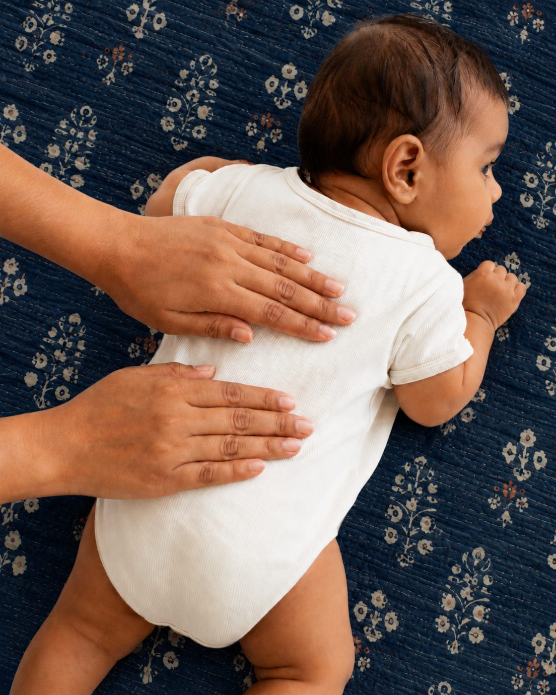 A parent's hands resting on a baby's back, the baby lying on an indigo block-print quilt.