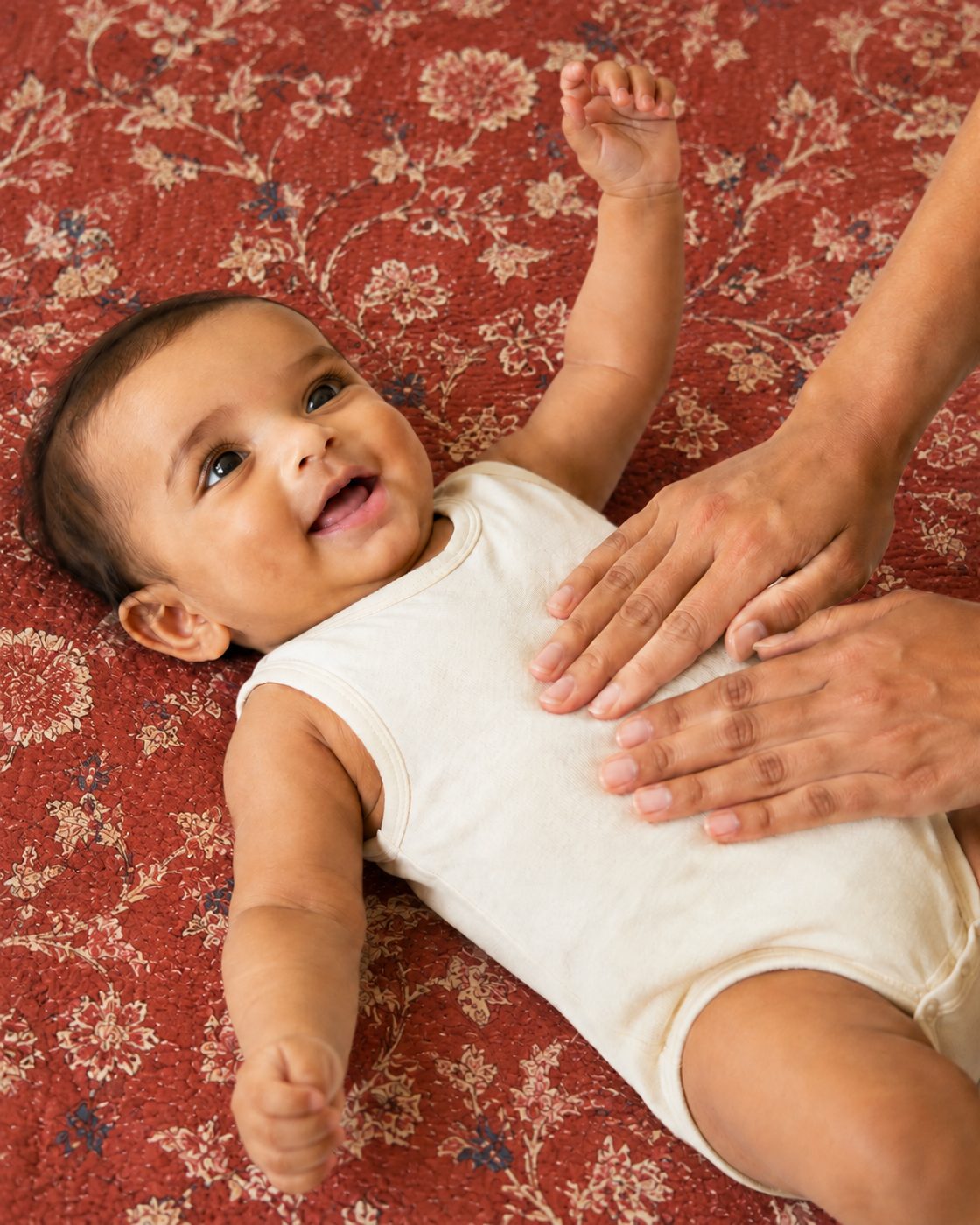 A smiling baby lying on a terracotta block-print quilt while a parent's hands rest on their chest.